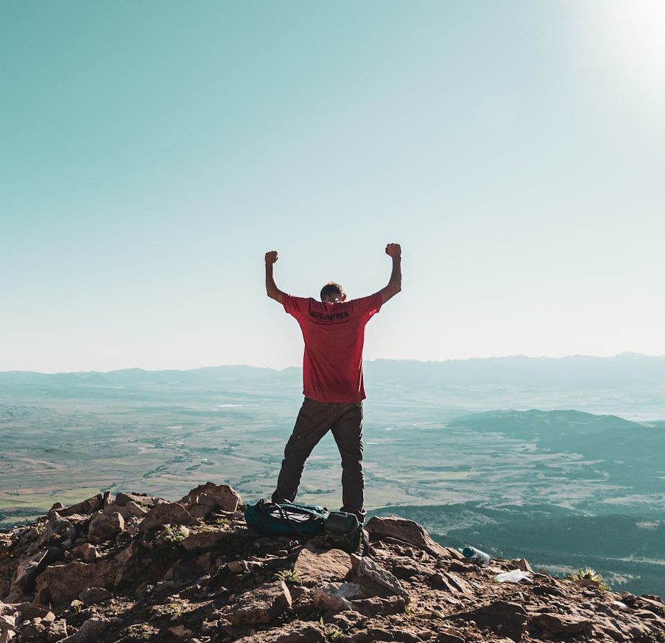 A man triumphantly celebrates on a rocky mountain summit, overlooking vast landscapes under clear skies.